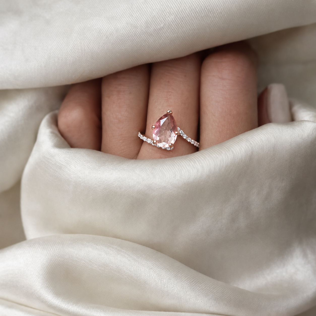 A close-up of a hand wearing a rose gold ring with a teardrop-shaped pink morganite gemstone and small clear accent stones, set against draped white satin fabric.