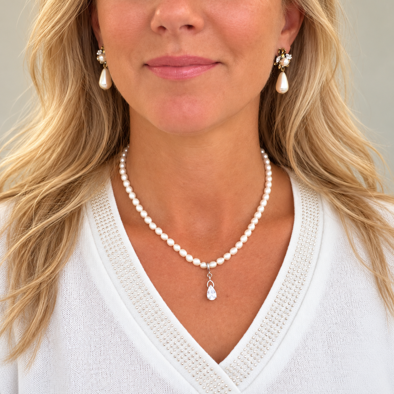 A close-up of a woman wearing a pearl necklace with a pear-shaped crystal pendant and matching pearl drop earrings with gold and pearl accents.