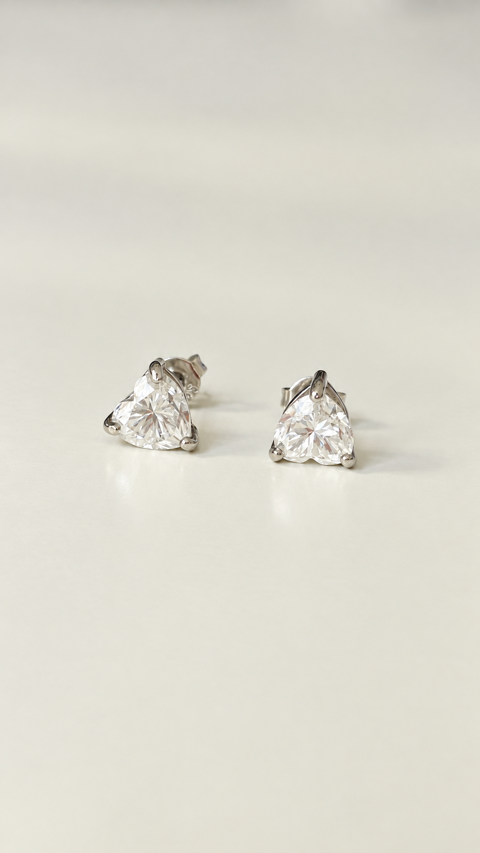 A pair of heart  shape moissanite stud earrings with clear faceted gemstones in a silver-toned setting, displayed on a light cream background.
