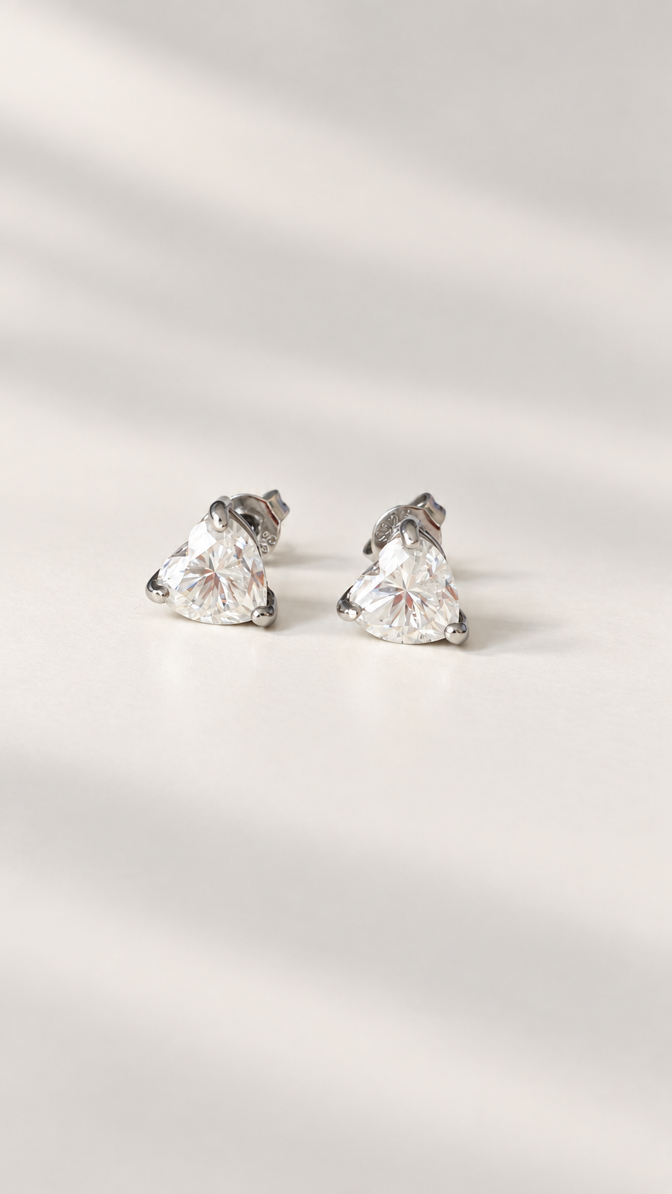 A pair of silver stud earrings with heart  shape moissanite gemstones, photographed on a light cream surface with soft diagonal shadows