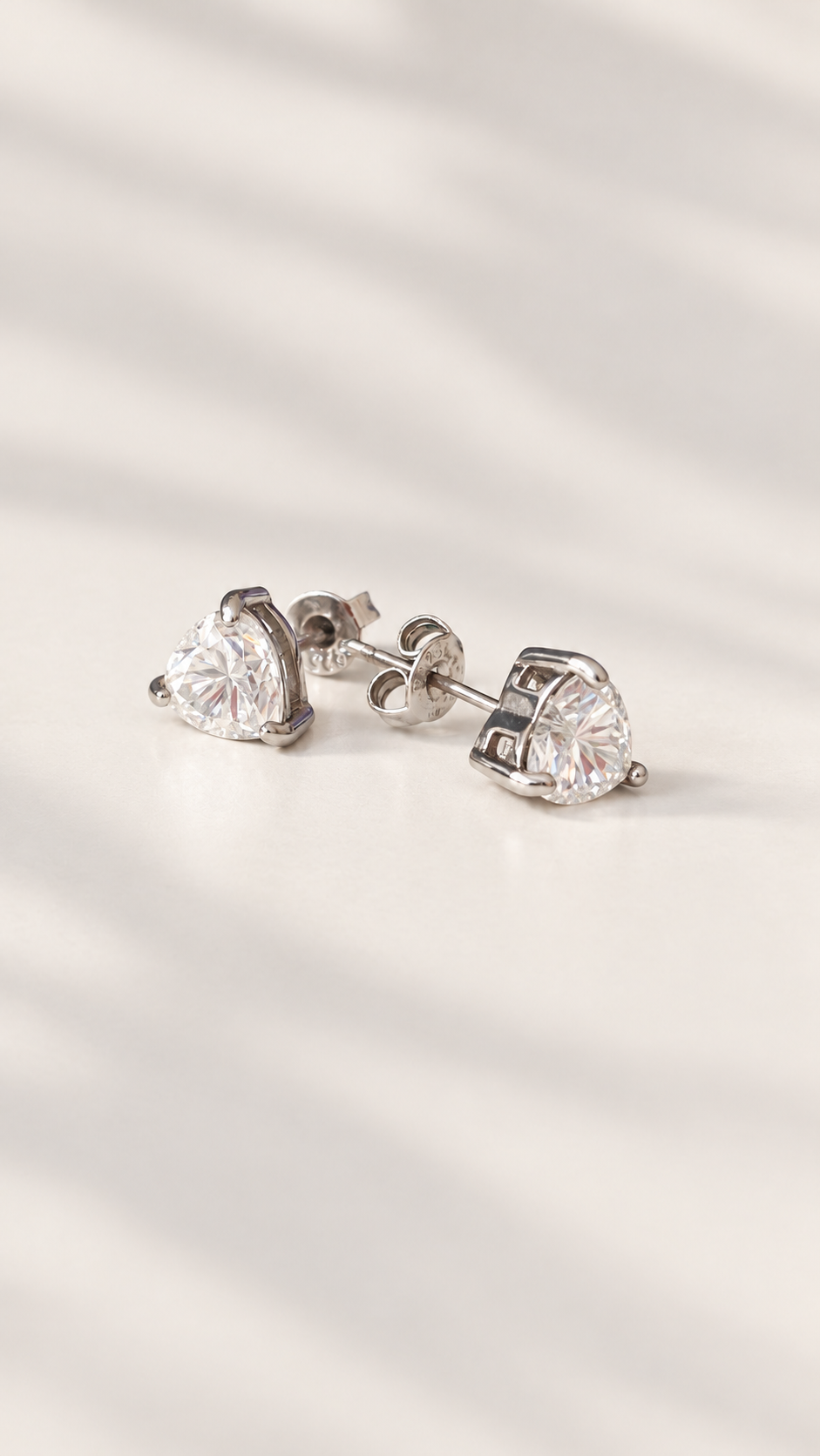 A pair of silver heart-shaped stud earrings with moissanite gemstones, displayed on a soft, light beige surface with elegant leaf shadows.
