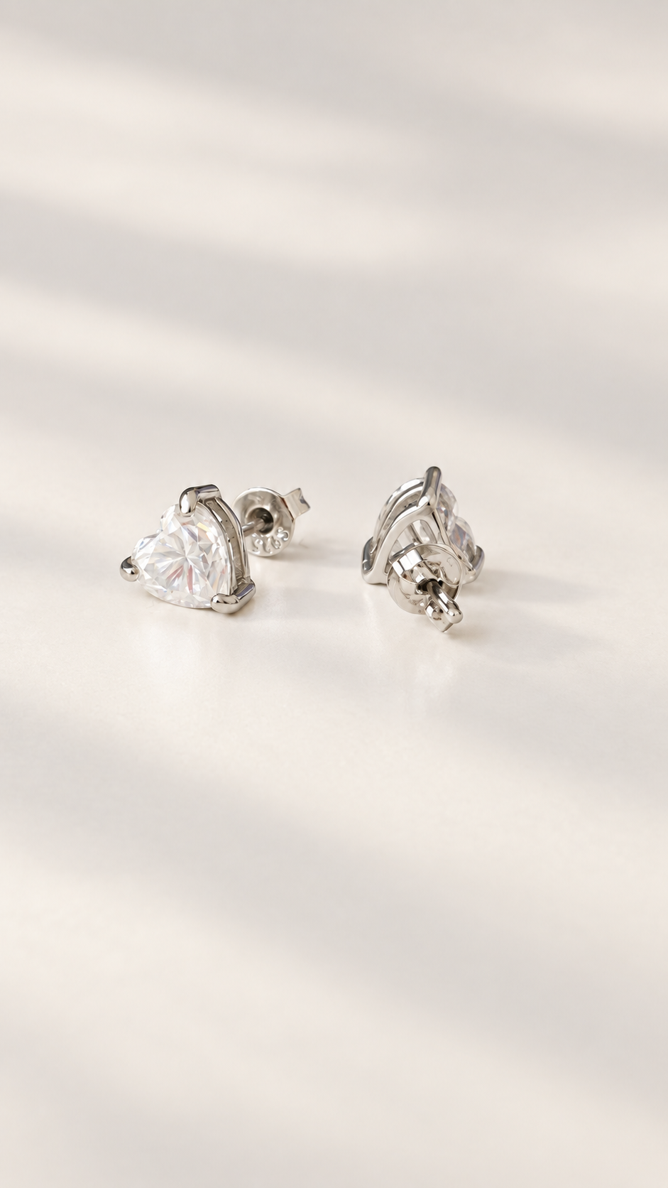 A pair of silver stud earrings with heart-shaped moissanite stones, displayed on a warm, softly lit cream surface.
