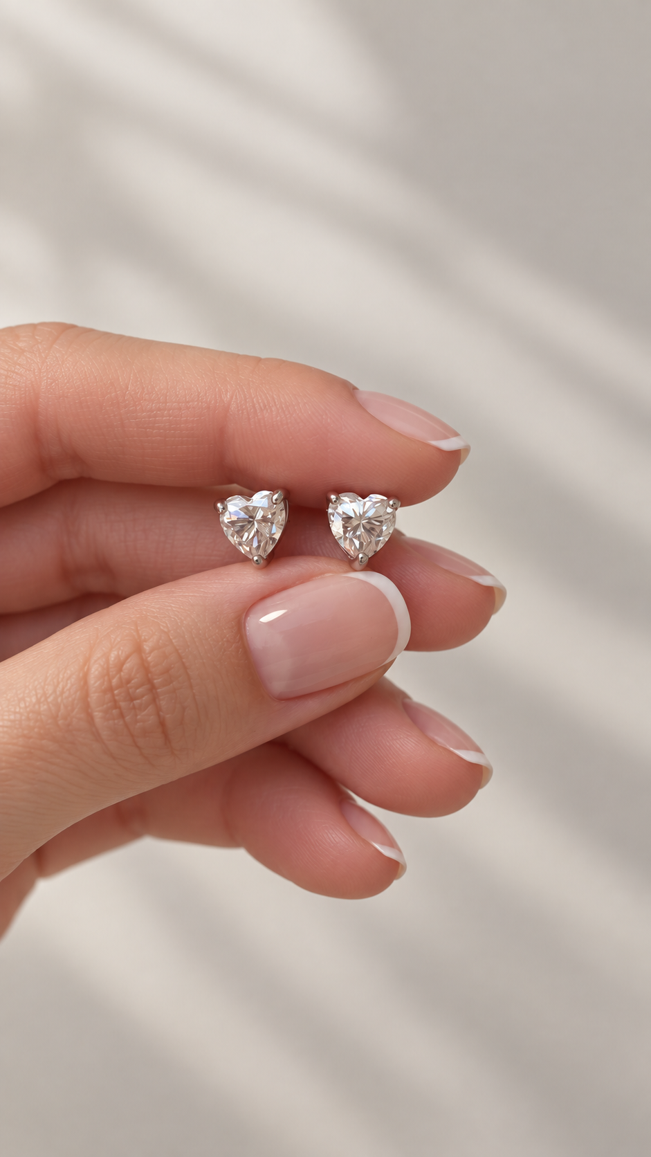 A close-up of a hand with a French manicure holding a pair of small heart-shaped stud earrings with sparkling moissanite gemstones in silver settings, against a soft, light gray background with diagonal shadows.