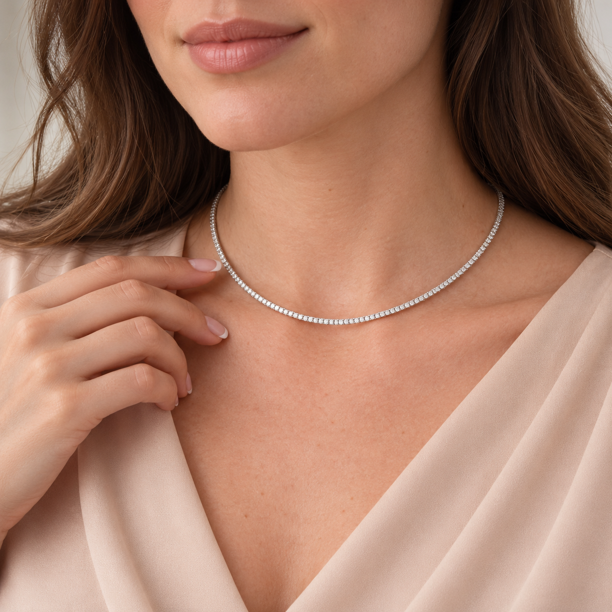 A close-up of a woman wearing a delicate silver tennis necklace with small round stones, paired with a light beige satin top.