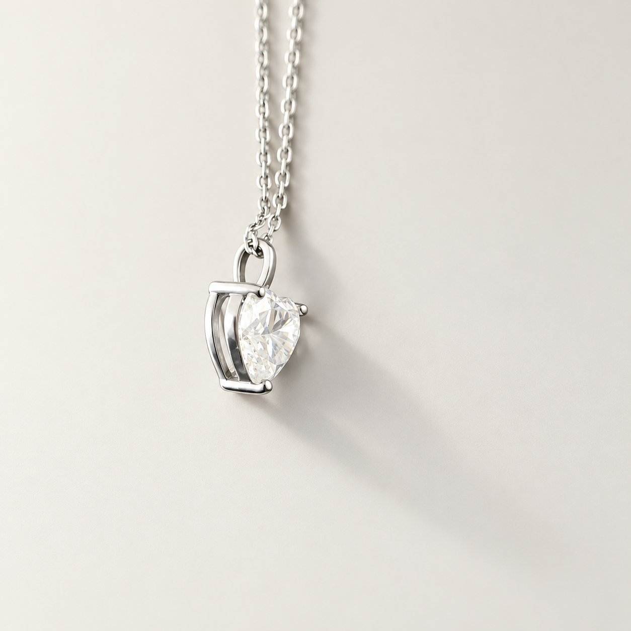 A silver pendant necklace featuring a heart-shaped clear moissanite gemstone in a prong setting, suspended from a delicate silver chain against a light beige background.
