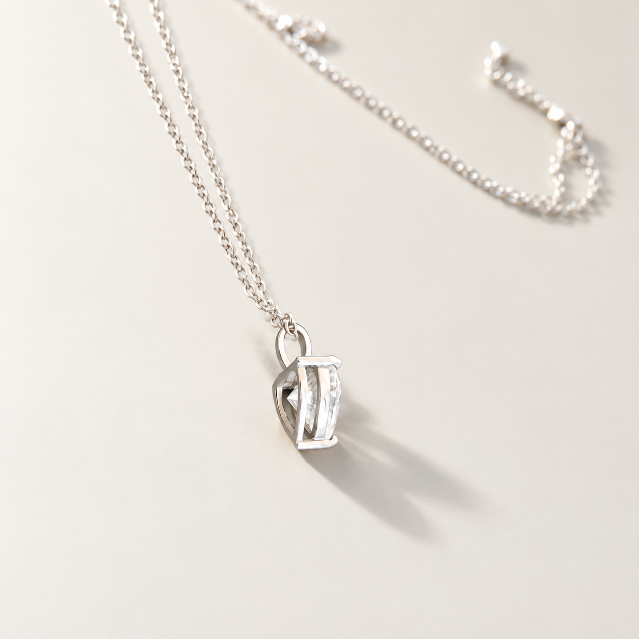 A silver chain necklace with a small openwork heart-shaped moissanite pendant, photographed on a clean light beige background with a soft shadow.