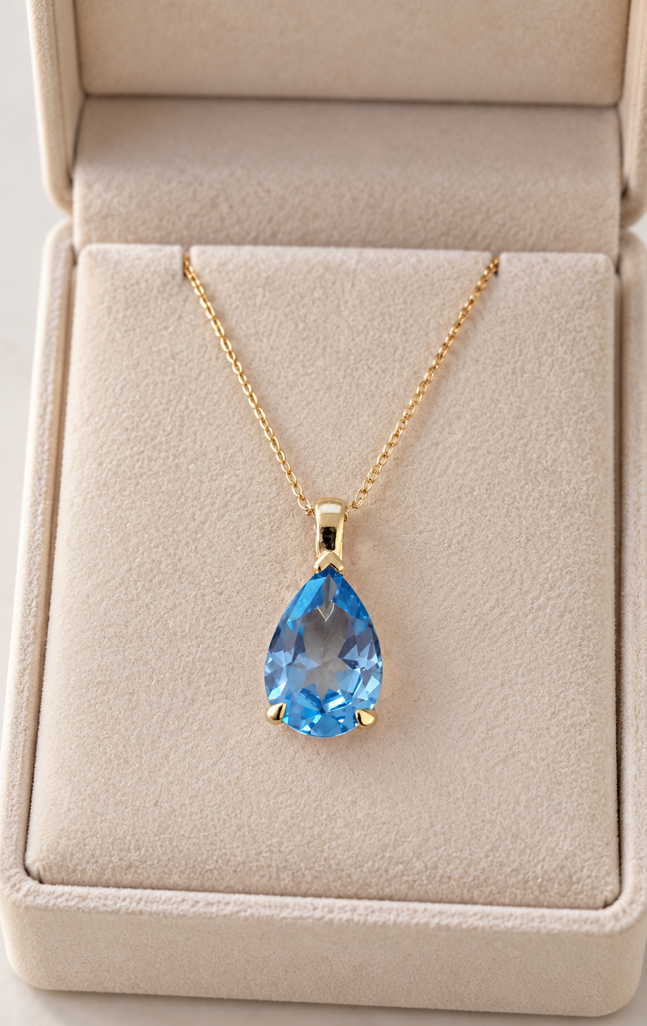 A gold pendant necklace with a teardrop-shaped light blue topaz gemstone, displayed inside an open beige jewelry box.