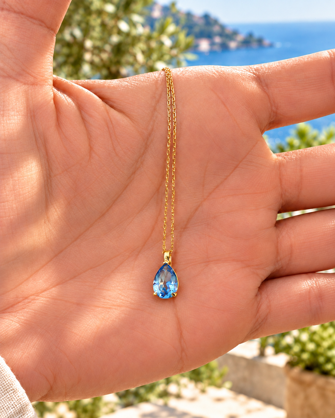 A gold chain necklace with a teardrop-shaped light blue topaz gemstone pendant, held in the palm of a hand with a blurred coastal background.