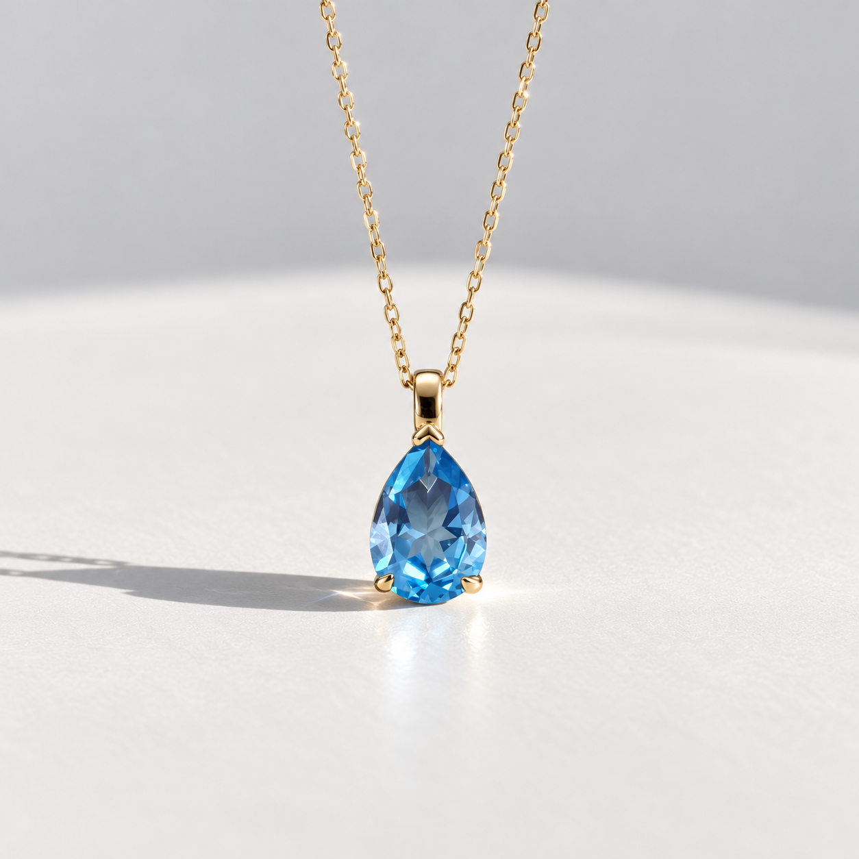 A gold chain necklace with a teardrop-shaped blue topaz gemstone pendant, displayed on a clean white surface with a soft shadow.