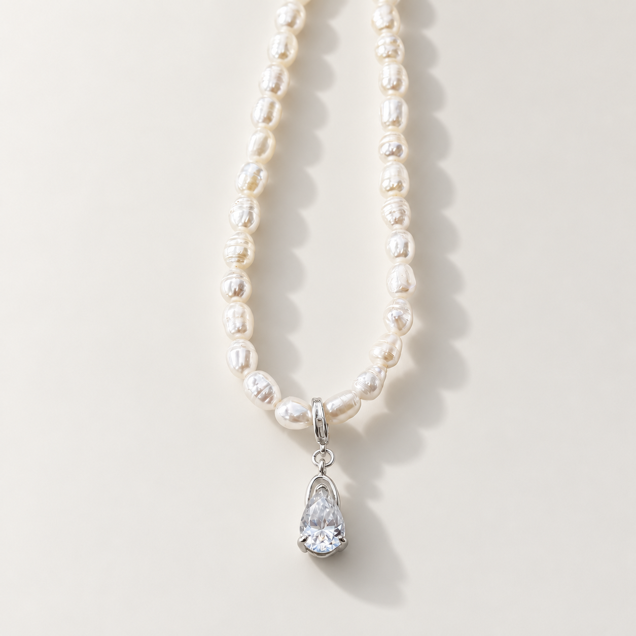 A strand of irregular white freshwater pearls with a silver-tone pendant featuring a teardrop-shaped clear moissanite gemstone, displayed on a light beige background.