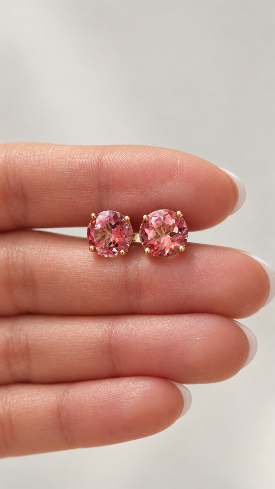 A pair of round pink topaz gemstone stud earrings with gold settings, held between a person's fingers against a soft light gray background.