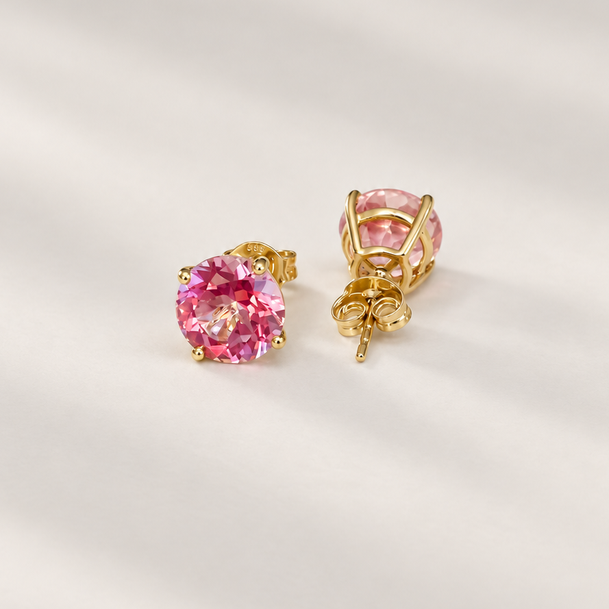 A pair of gold stud earrings with round pink topaz gemstones, shown on a soft, light beige fabric background.