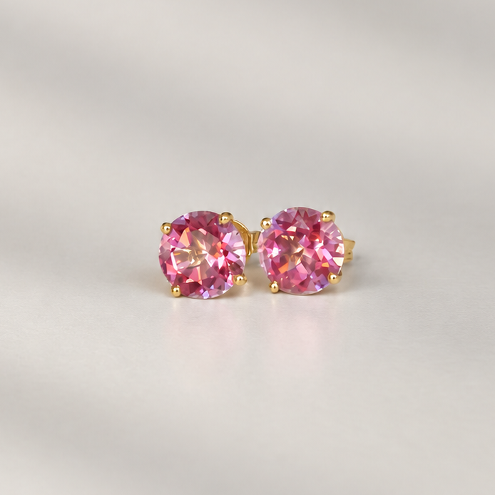 A pair of gold stud earrings with round pink topaz gemstones, displayed on a light gray background with soft diagonal shadows.