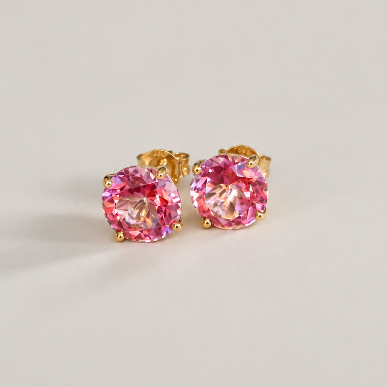 A pair of gold stud earrings with round-cut pink topaz gemstones, displayed on a clean, light beige background.