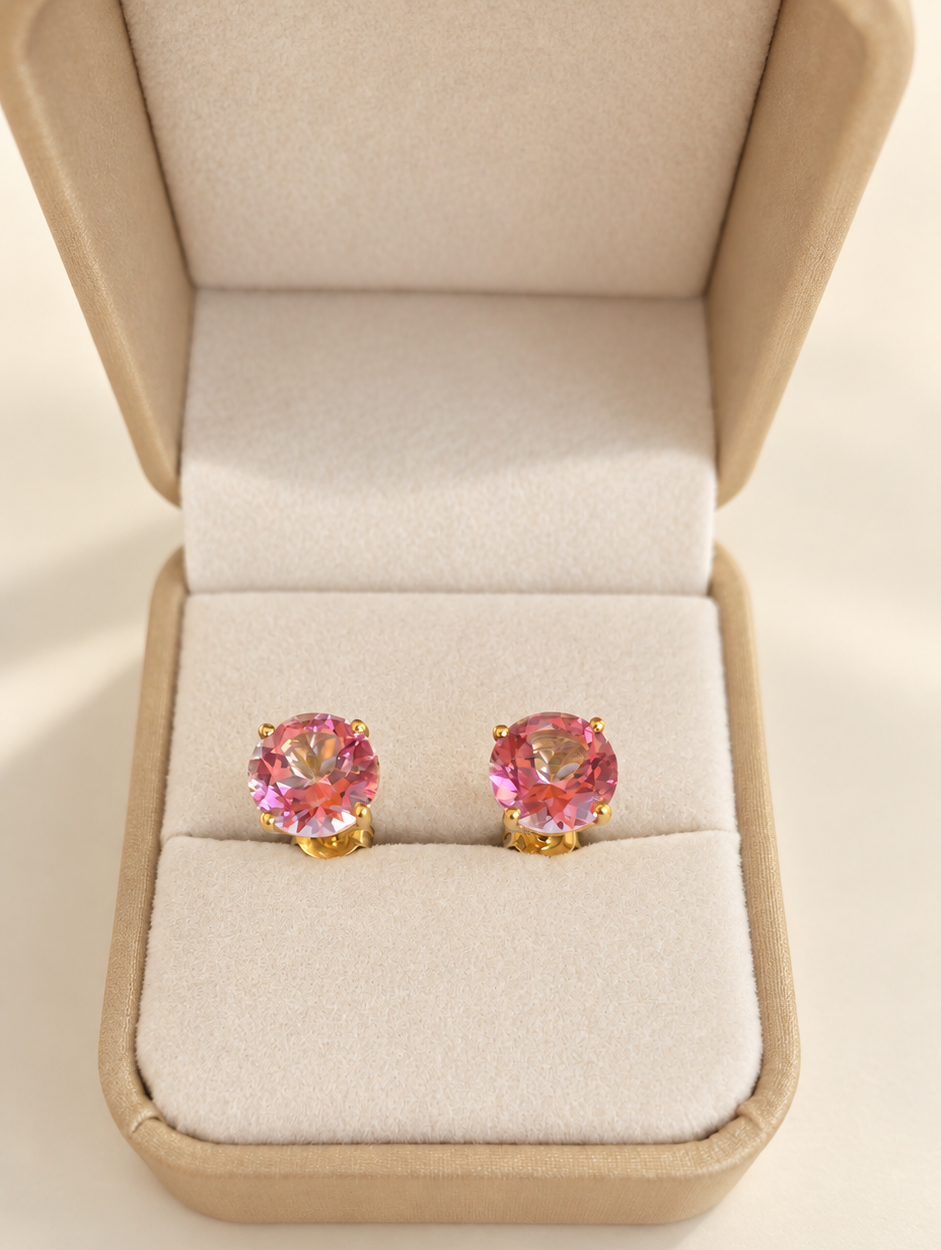 A pair of gold stud earrings with round pink topaz gemstones, presented in an open beige jewelry box on a light beige background.