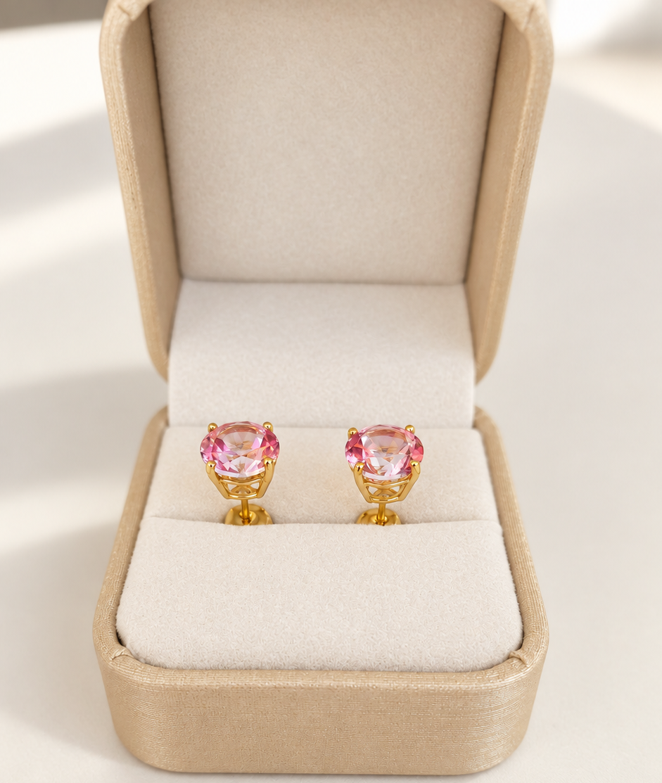 A pair of gold stud earrings with heart-shaped pink topaz gemstones, presented in an open beige jewelry box on a light cream background.