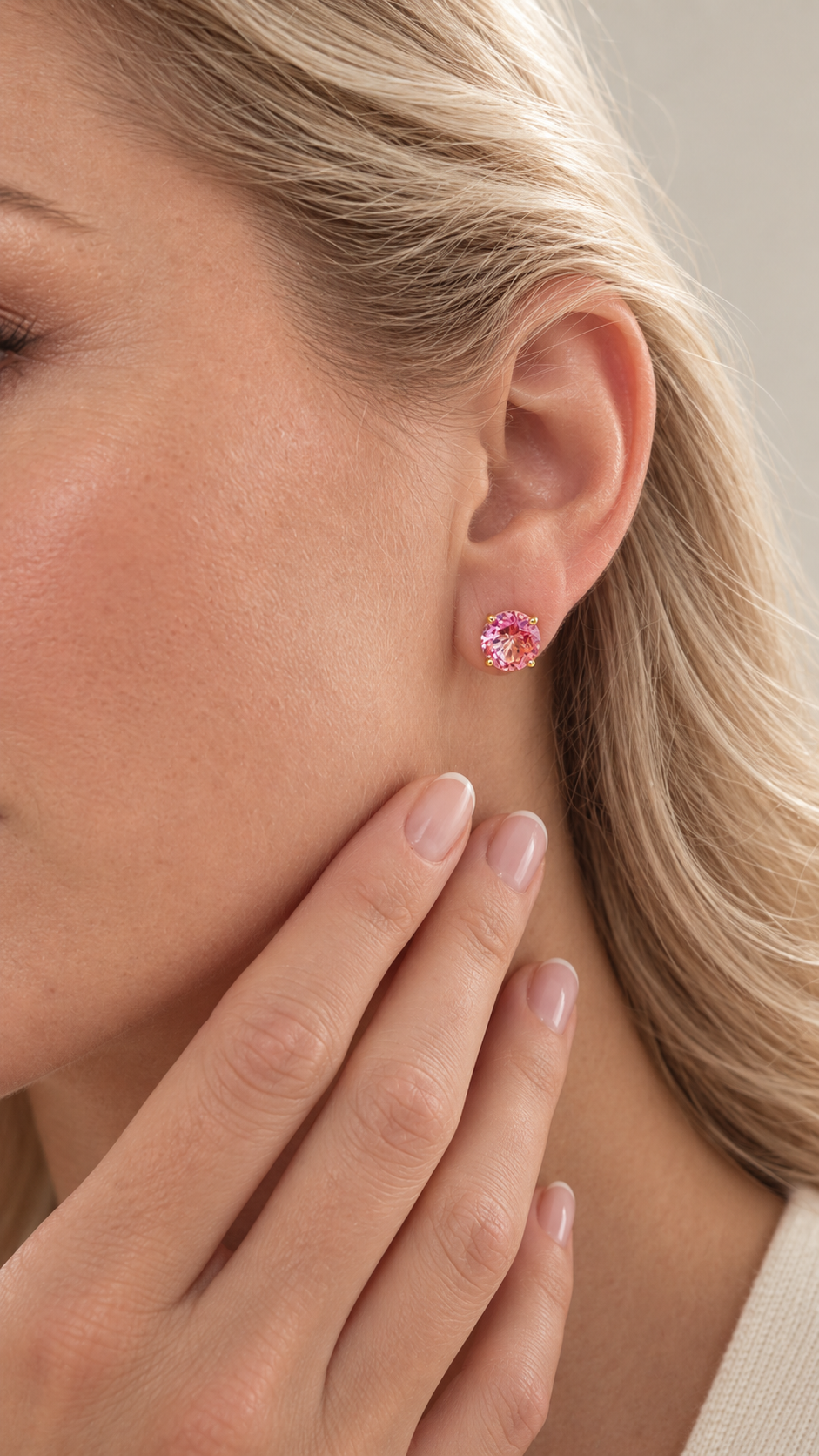 A close-up of a woman's ear wearing a small round stud earring with a pink topaz gemstone and a gold-toned setting, with her hand resting near her face.