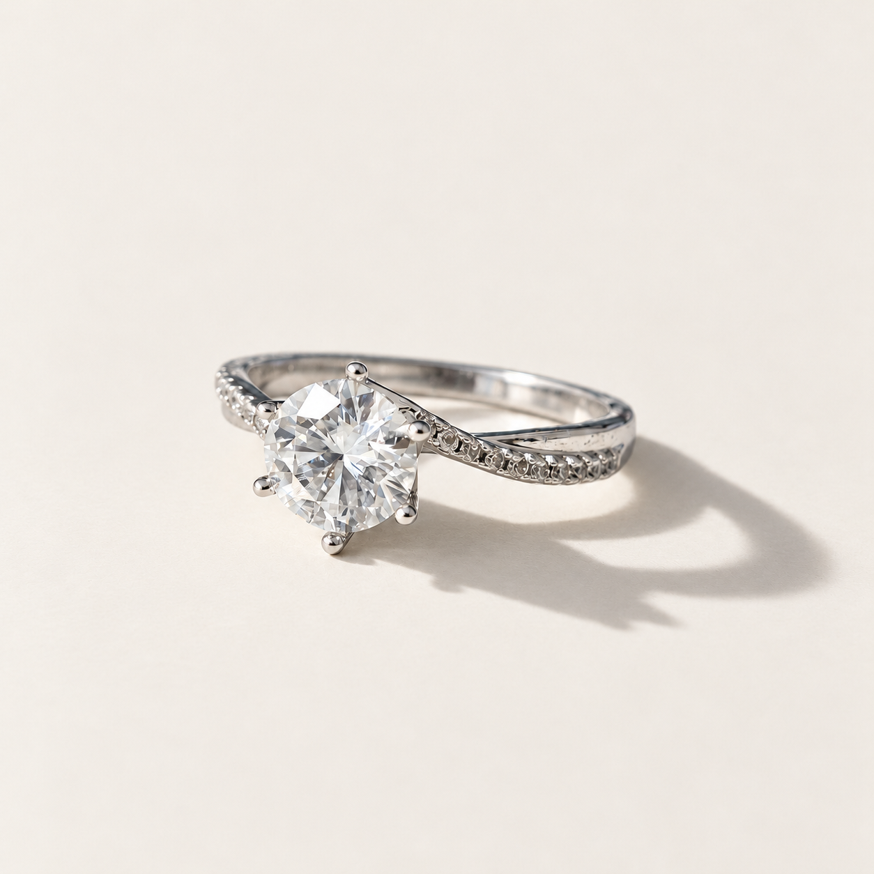 A silver solitaire engagement ring with a round-cut clear moissanite center stone and a pavé-set diamond band, photographed on a warm off-white background with a soft shadow.