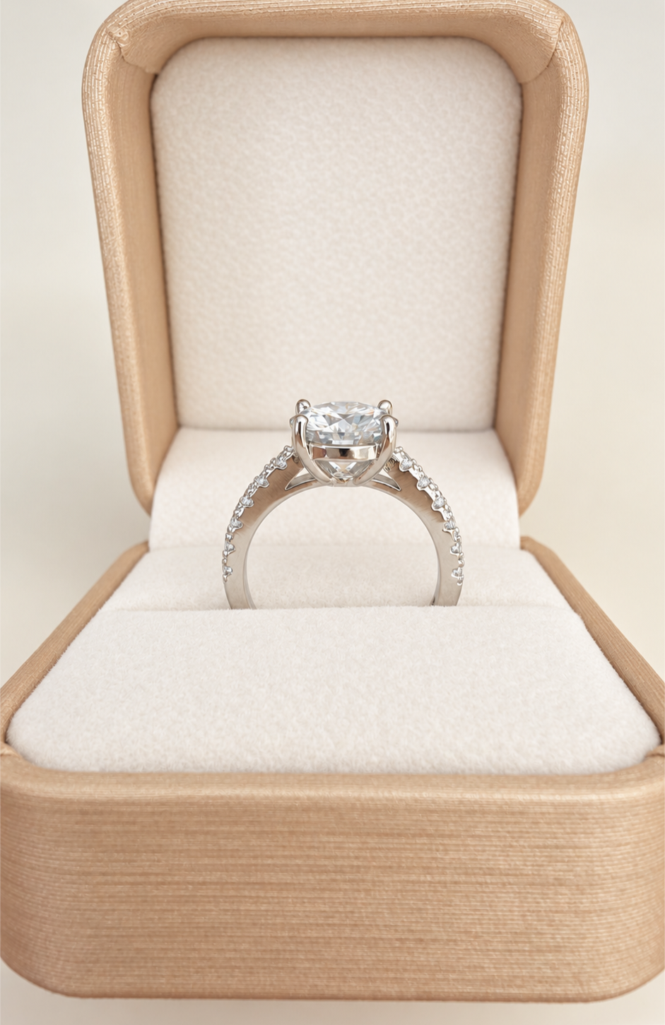 A silver-toned solitaire engagement ring with a round-cut center moissanite stone and small pavé accents on the band, displayed inside an open champagne-colored jewelry box with a cream velvet interior.