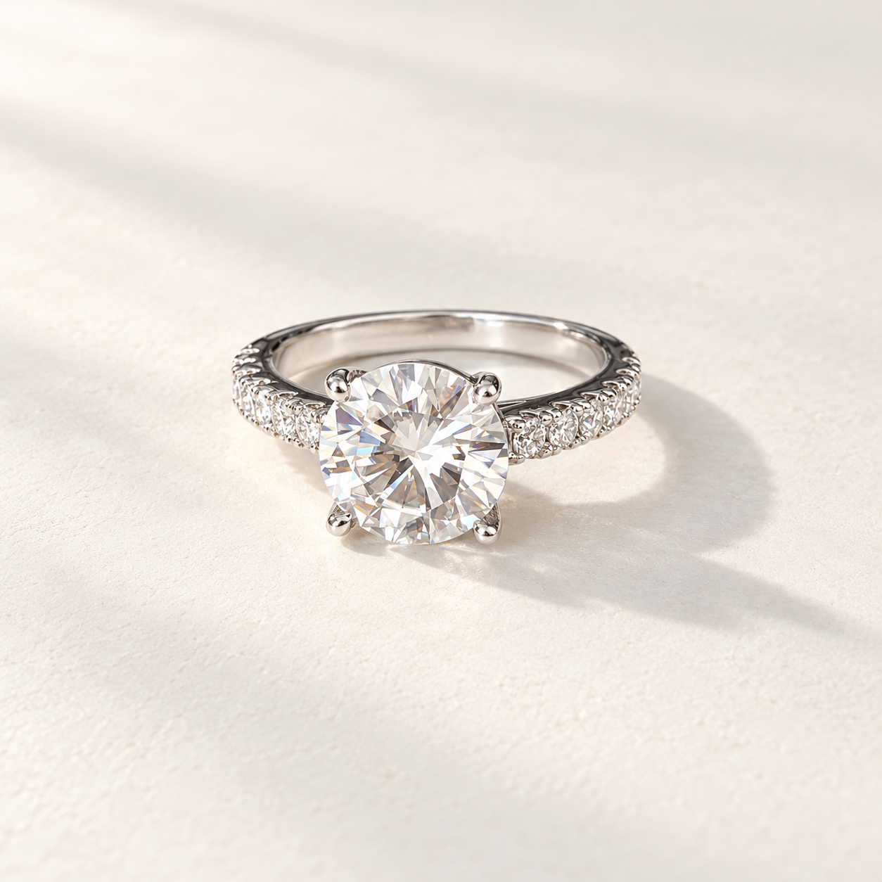 A round-cut moissanite solitaire engagement ring with a pavé band, set in a silver-colored metal on a textured off-white background with soft shadows.