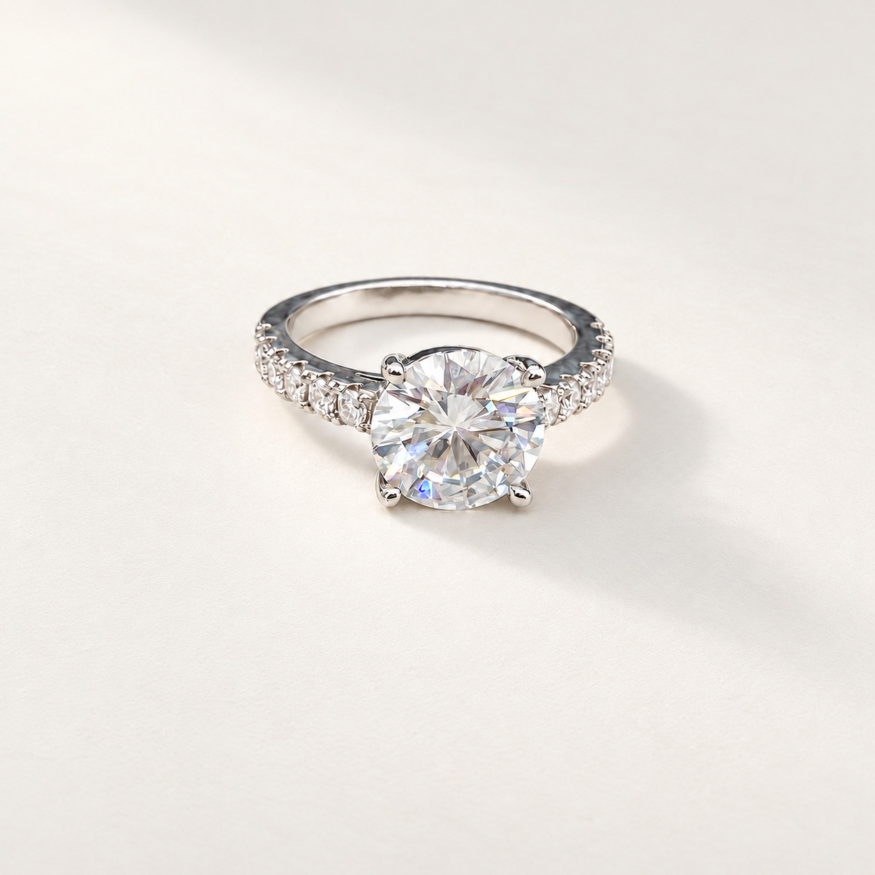 A silver solitaire engagement ring with a round brilliant-cut center moissanite stone and a pavé-set band, photographed on a clean, light beige background.