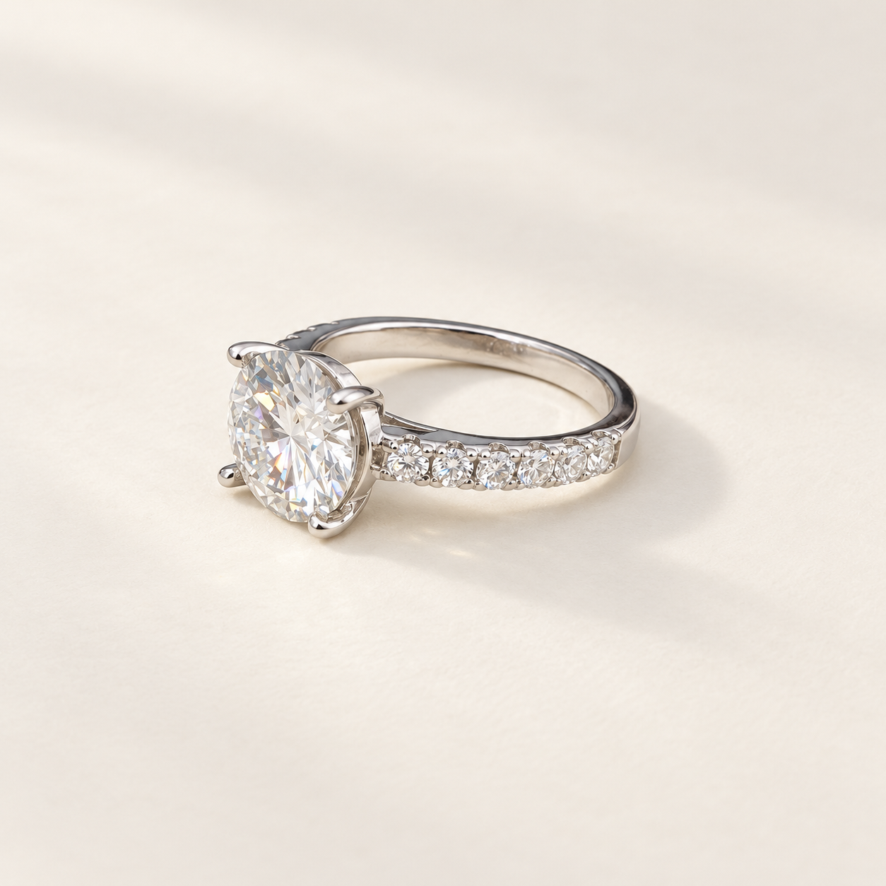 A silver solitaire engagement ring with a round brilliant-cut moissanite center stone and a pavé-set diamond band, photographed on a clean, light beige textured background.