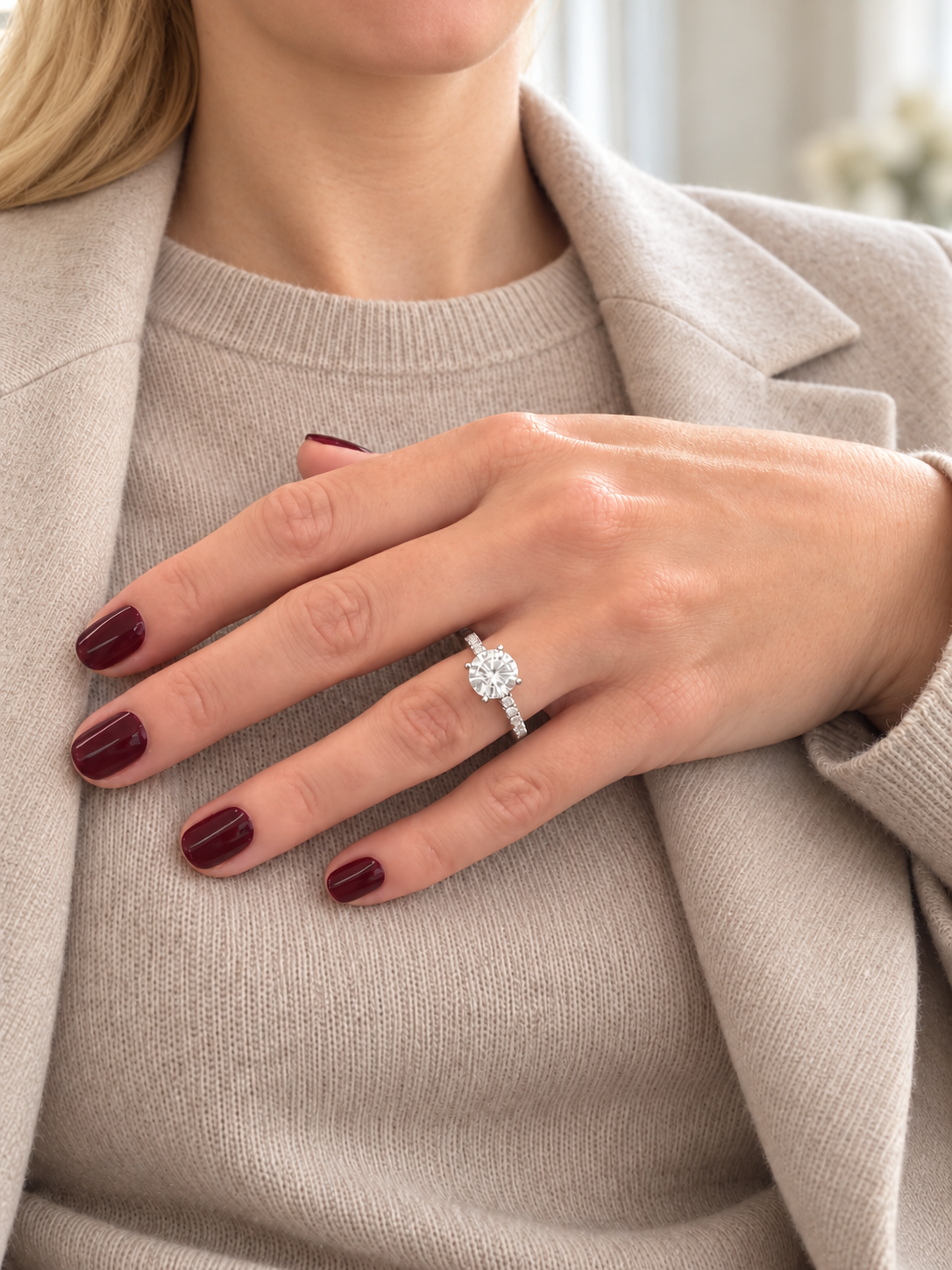 A close-up lifestyle shot of a woman wearing a diamond engagement ring on her ring finger, styled with a beige knit sweater and a light beige blazer.