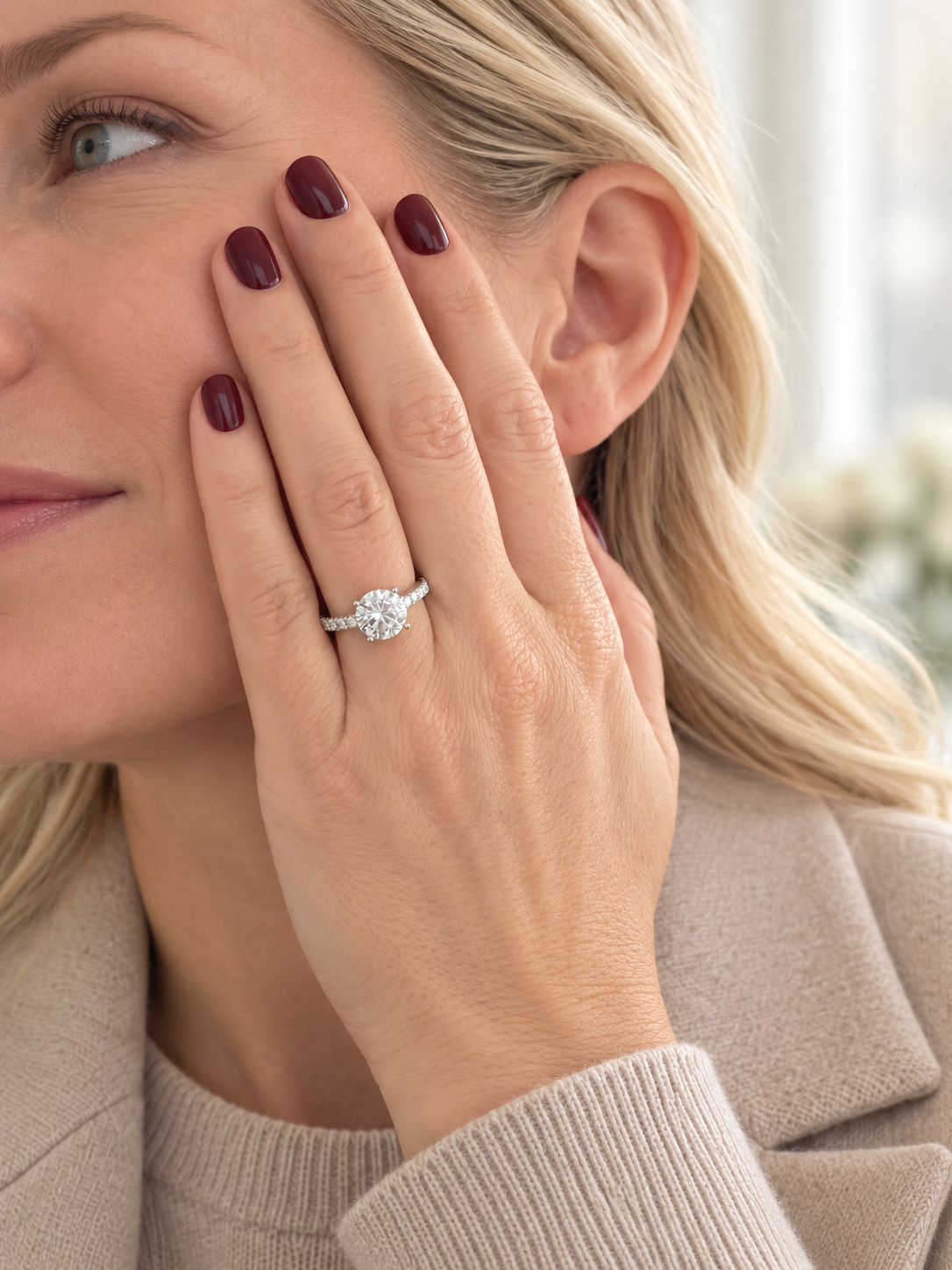A close-up lifestyle shot of a woman wearing a silver diamond engagement ring on her finger, with her hand resting against her cheek. She has blonde hair and dark burgundy nail polish, wearing a beige knit sweater and a beige coat.