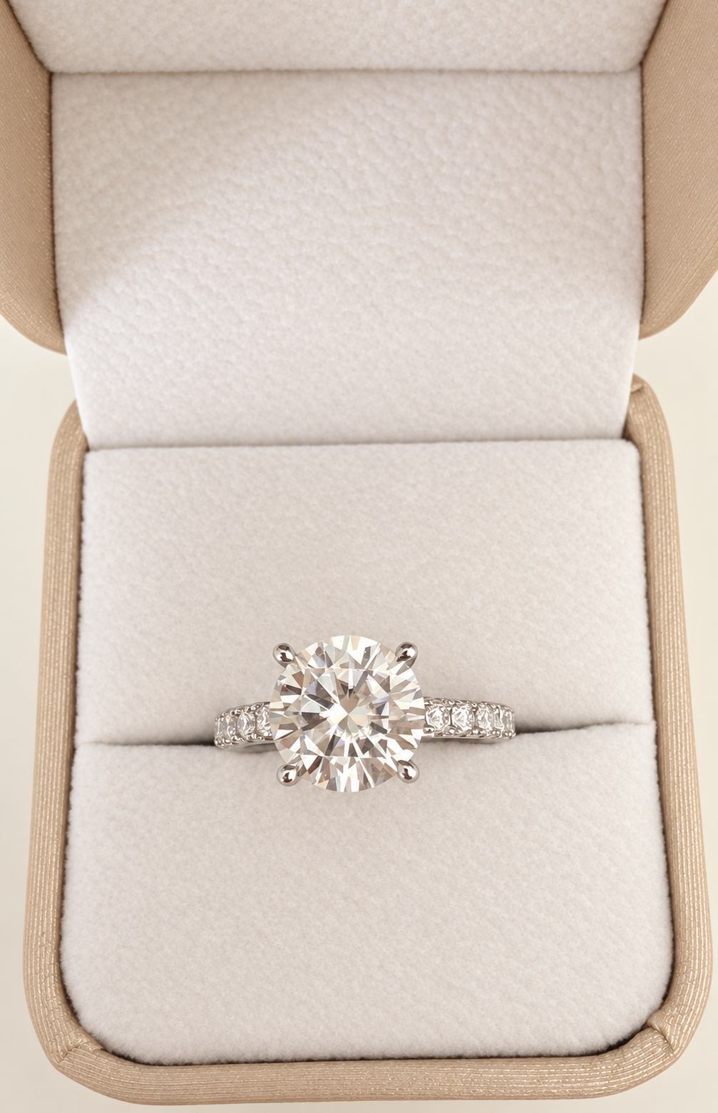 A round-cut moissanite diamond engagement ring with a silver-toned band set with small diamonds, displayed in an open beige jewelry box with a cream-colored velvet interior.