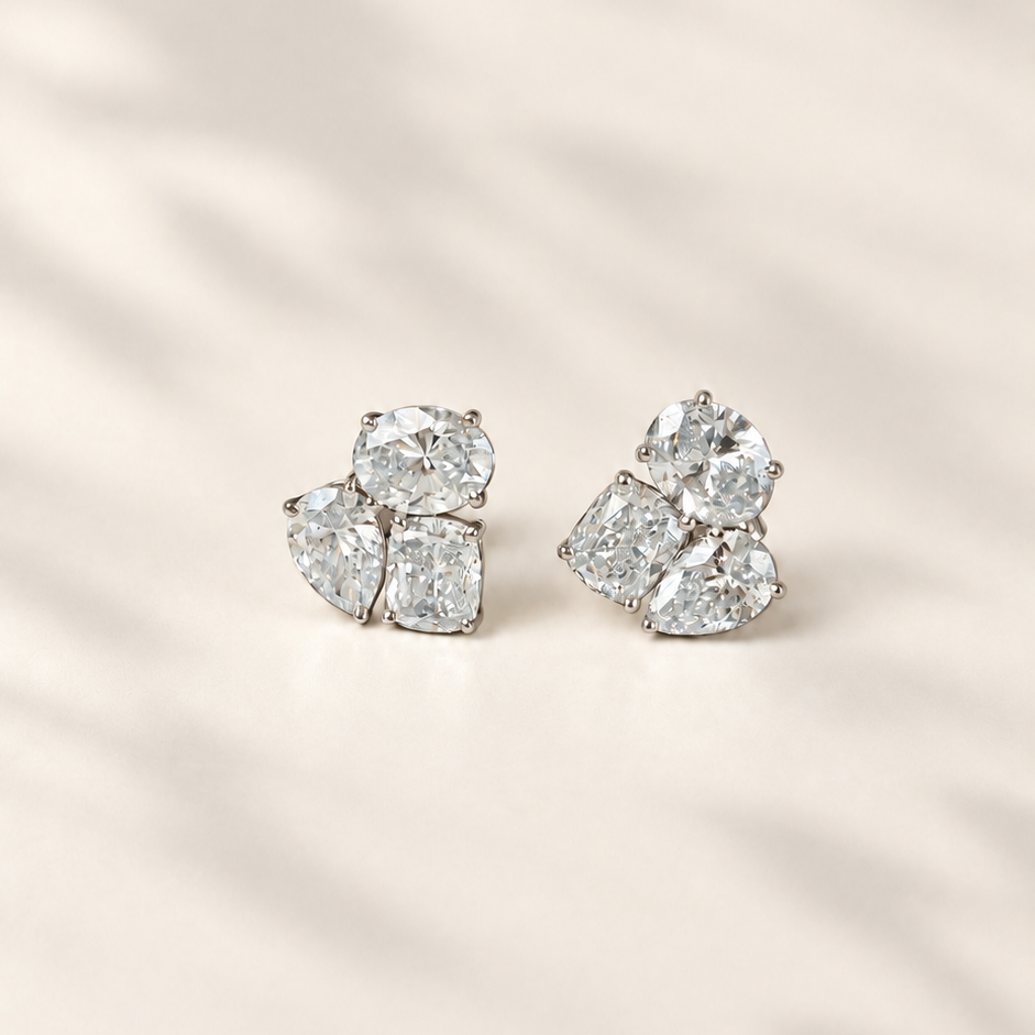 A pair of diamond stud earrings with a cluster design, featuring pear and cushion-cut diamonds in a silver-toned setting, displayed on a warm beige surface with soft leaf shadows.